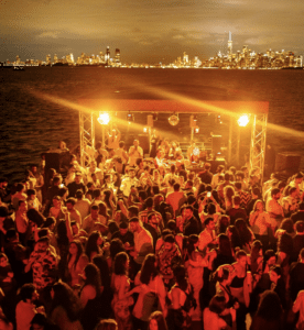 A large crowd dances at an outdoor night party on a waterfront, energized by New York music, with city skyline lights and tall buildings visible across the water.